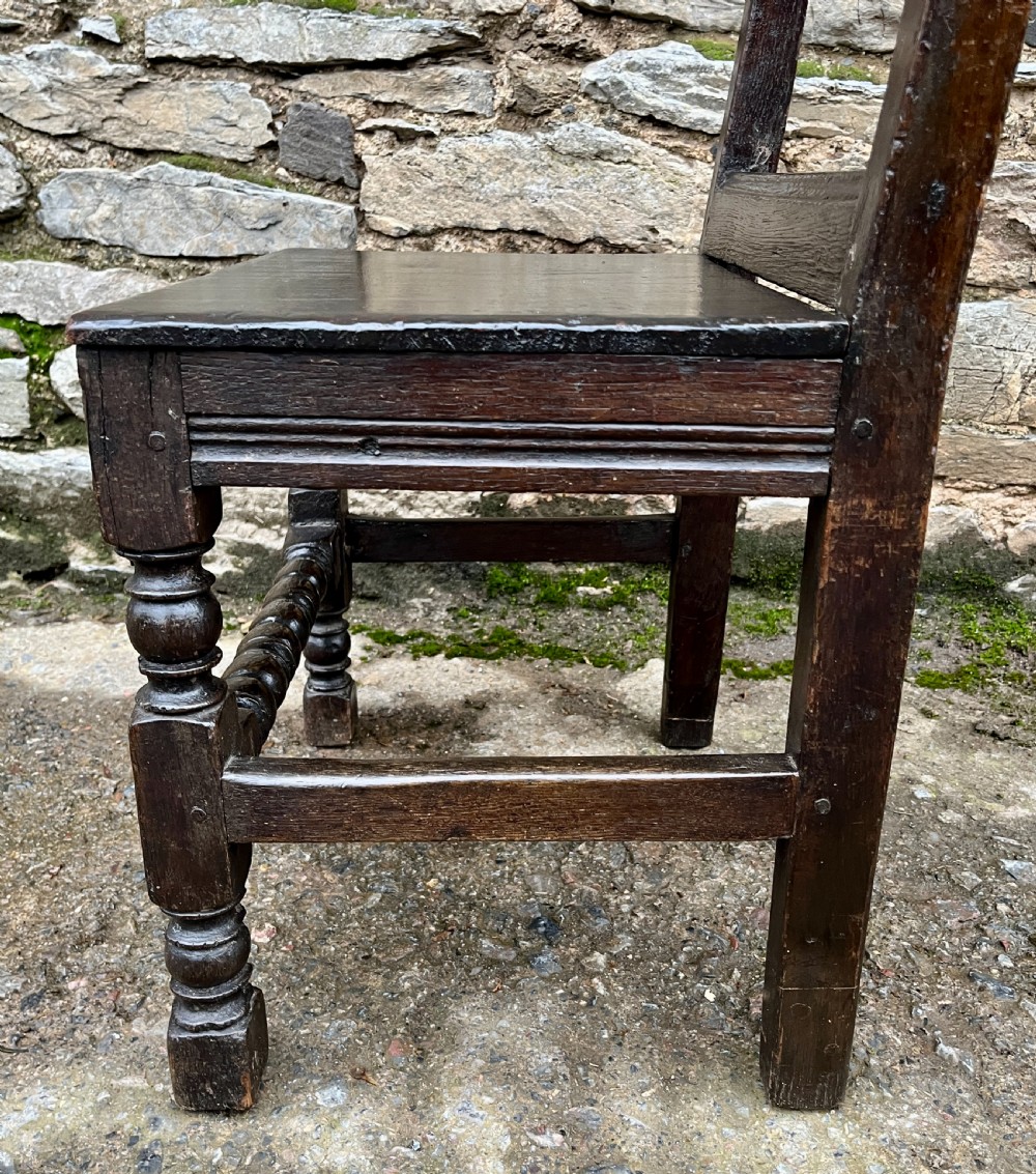 A dark oak 17th-century backstool featuring a carved crested top rail, solid wooden seat, turned front legs, and a stretchered base. View X