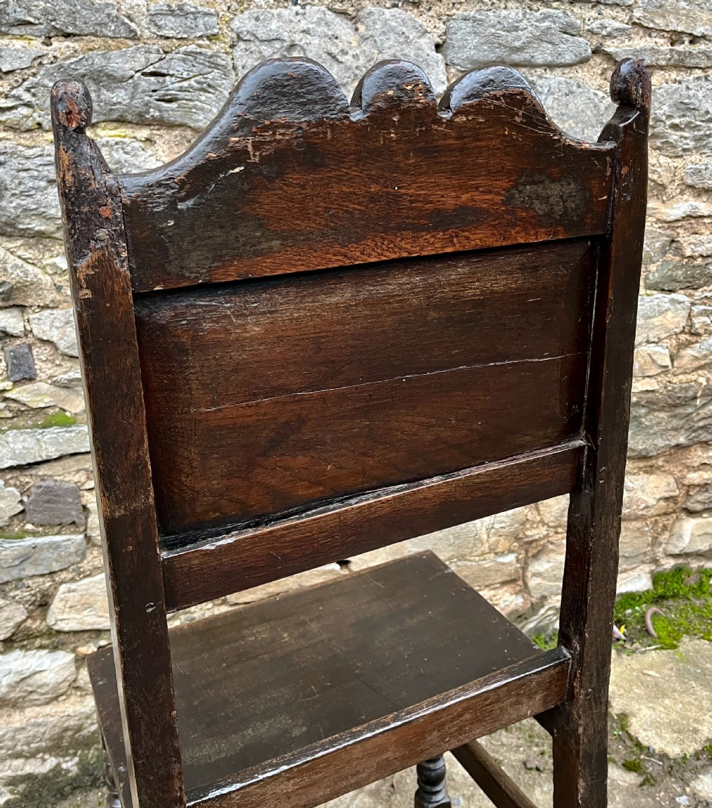 A dark oak 17th-century backstool featuring a carved crested top rail, solid wooden seat, turned front legs, and a stretchered base. View XI