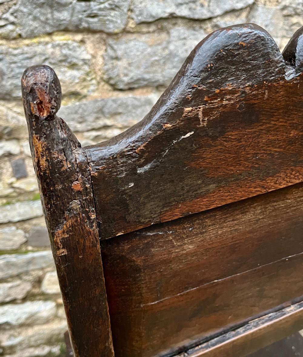 A dark oak 17th-century backstool featuring a carved crested top rail, solid wooden seat, turned front legs, and a stretchered base. View XII