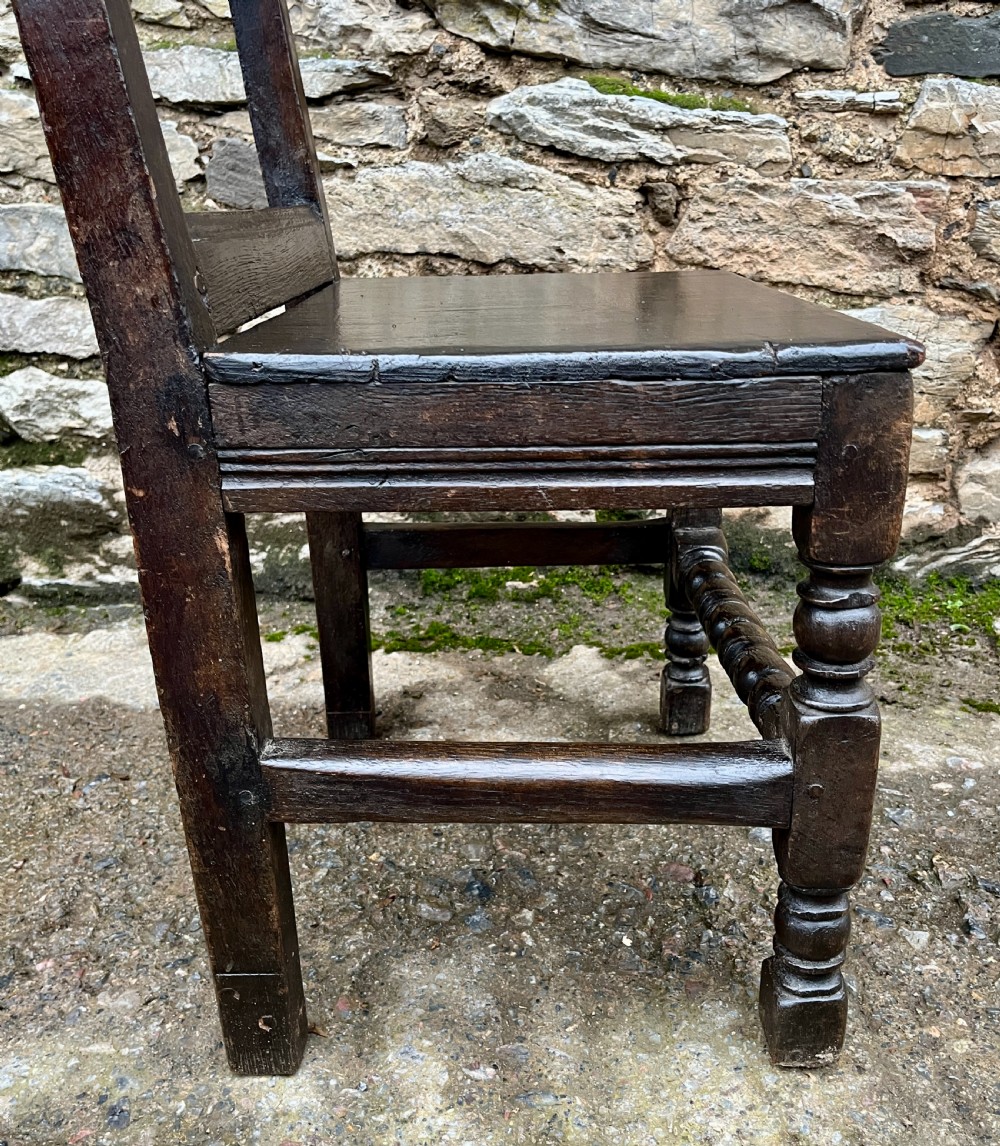 A dark oak 17th-century backstool featuring a carved crested top rail, solid wooden seat, turned front legs, and a stretchered base. View XIV