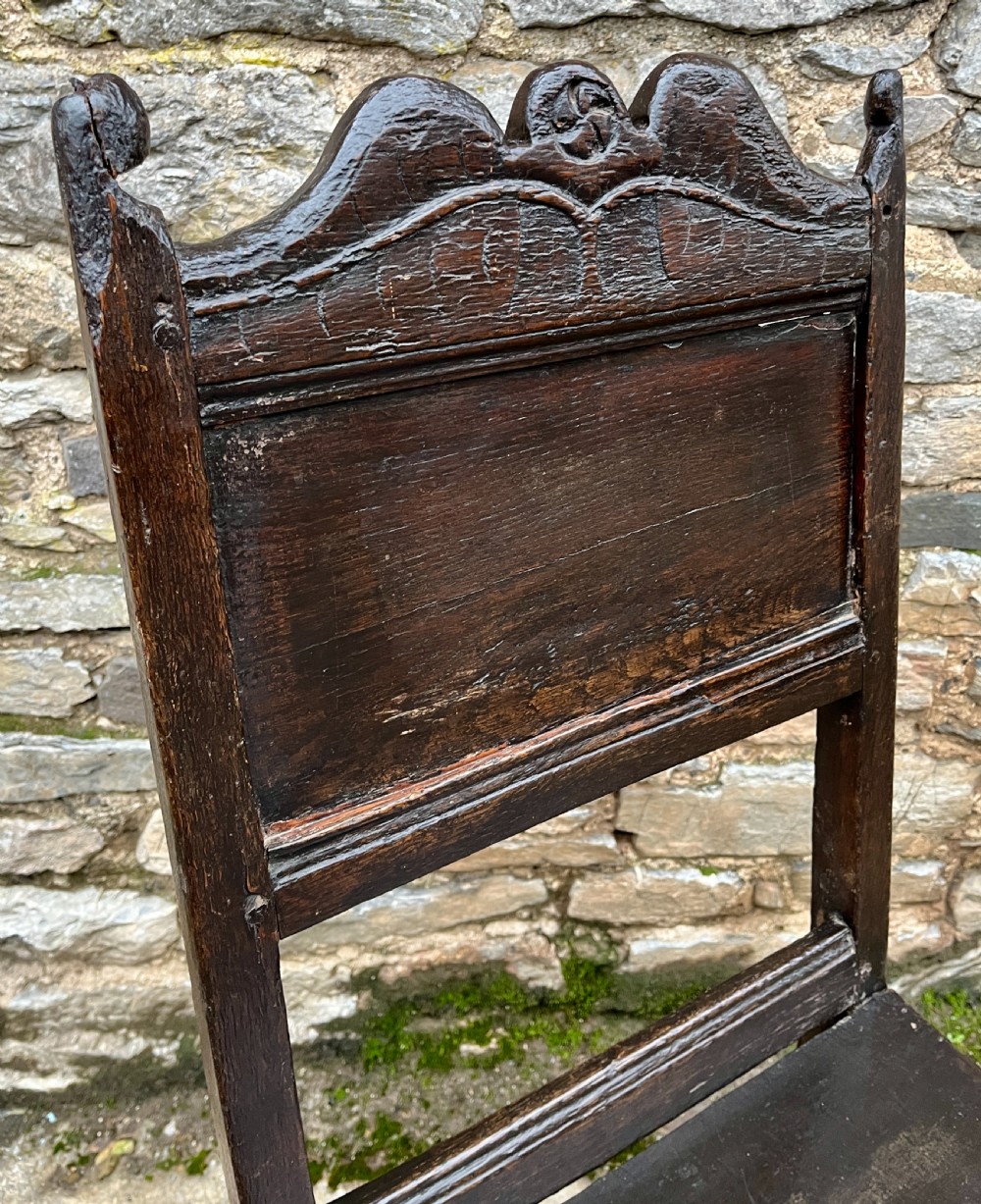 A dark oak 17th-century backstool featuring a carved crested top rail, solid wooden seat, turned front legs, and a stretchered base. View XV