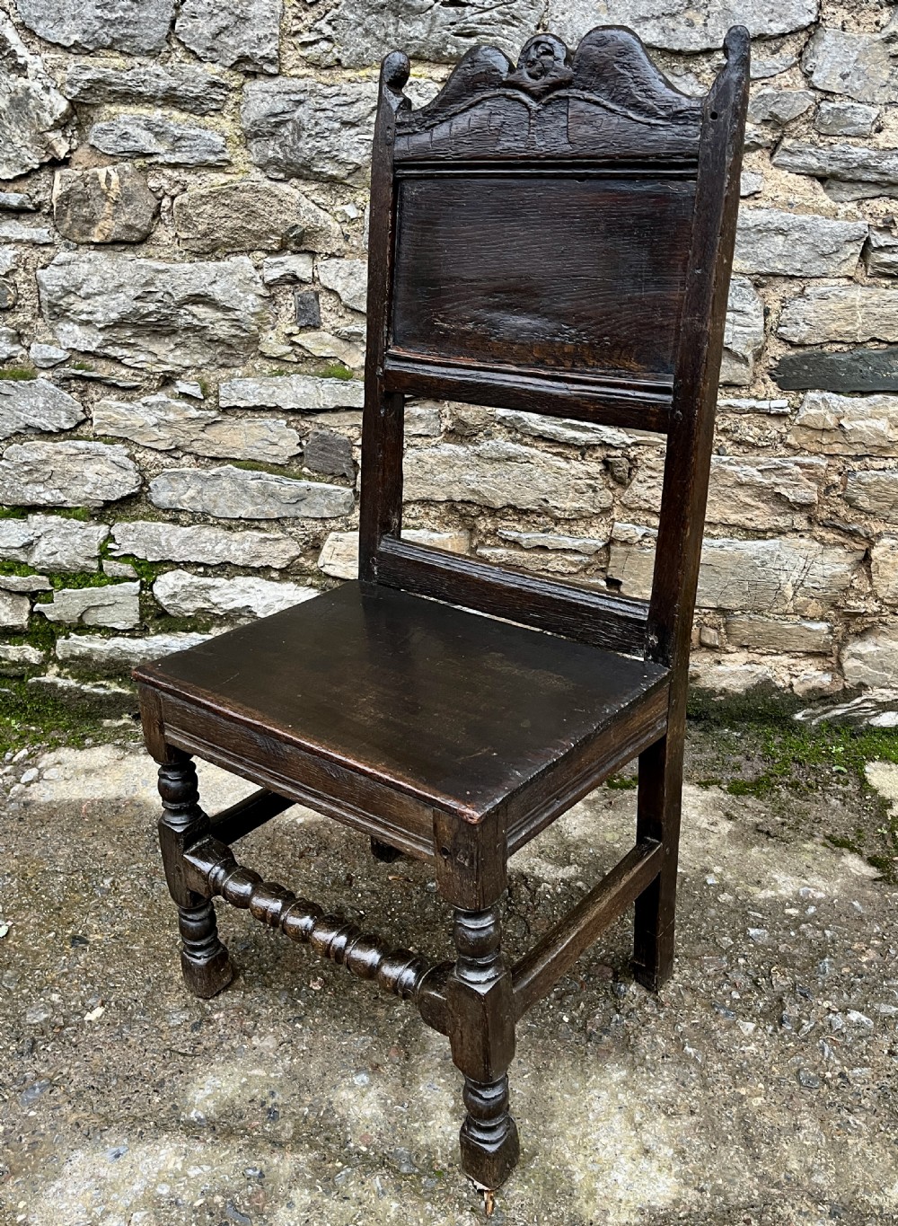 A dark oak 17th-century backstool featuring a carved crested top rail, solid wooden seat, turned front legs, and a stretchered base. View XVI