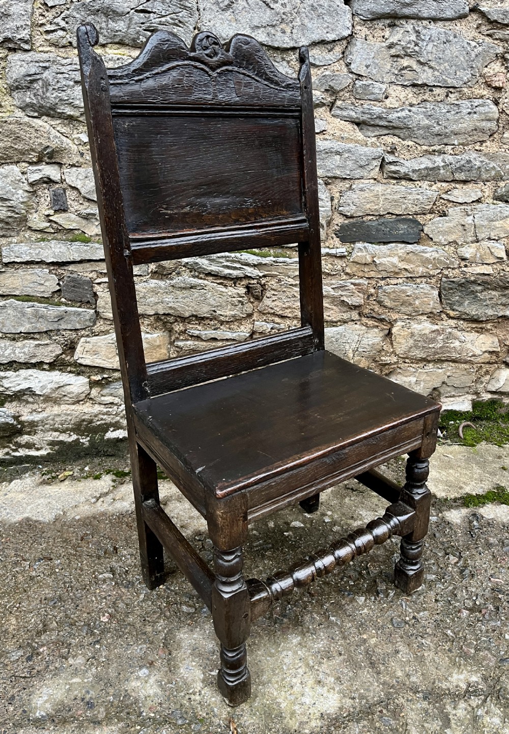A dark oak 17th-century backstool featuring a carved crested top rail, solid wooden seat, turned front legs, and a stretchered base. View XVIII