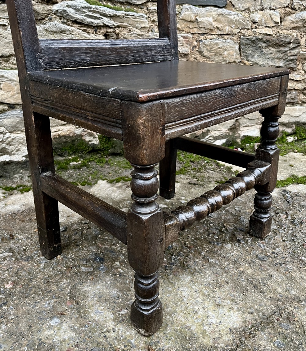A dark oak 17th-century backstool featuring a carved crested top rail, solid wooden seat, turned front legs, and a stretchered base. View XIX