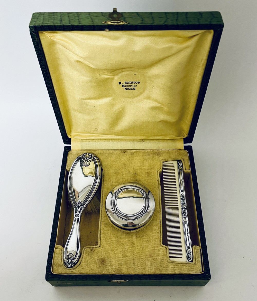 An open green textured box containing a French silver vanity set with a silver hairbrush, comb, and round lidded jar set into a yellow silk lining. View II