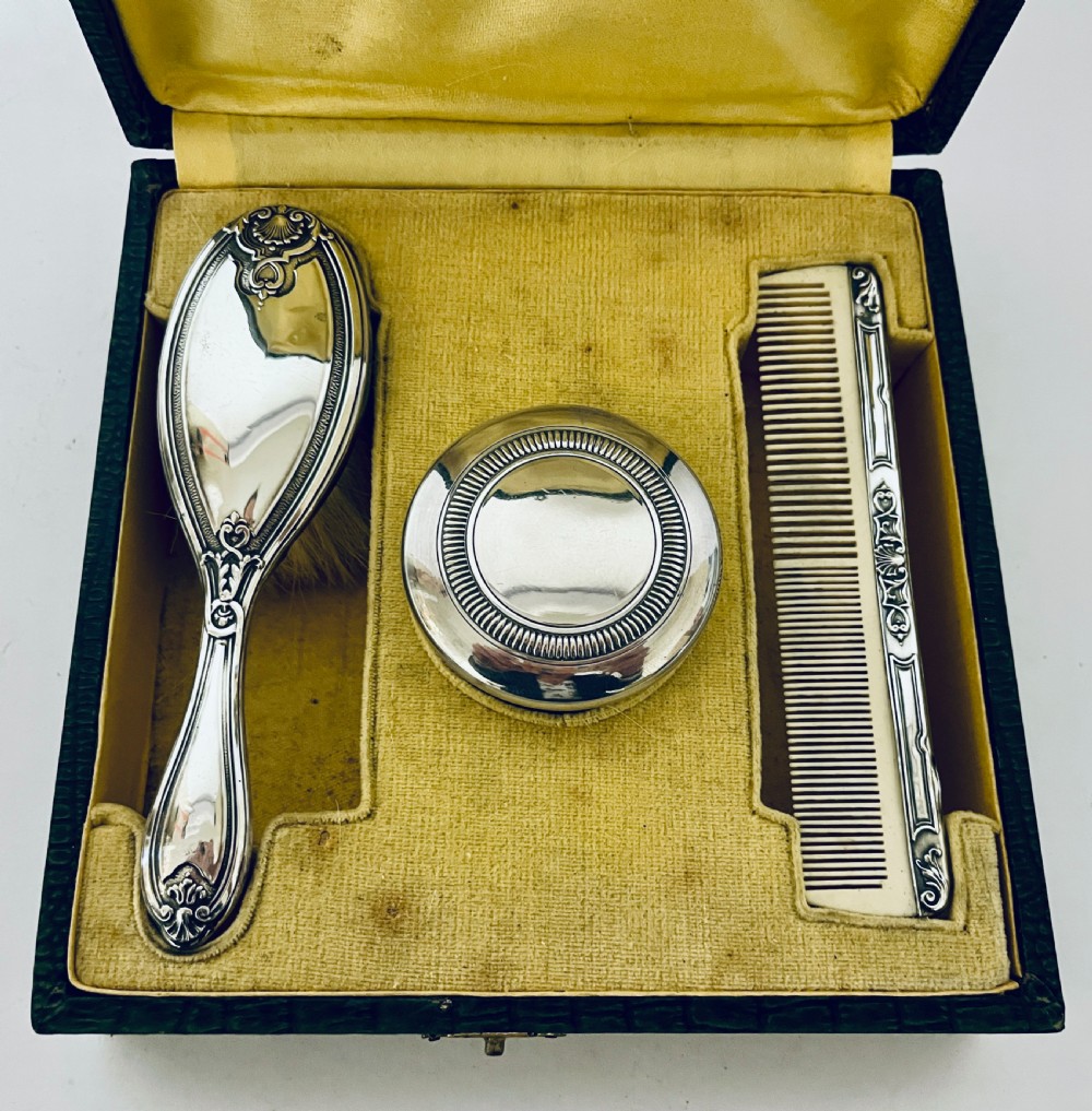 An open green textured box containing a French silver vanity set with a silver hairbrush, comb, and round lidded jar set into a yellow silk lining. View IV