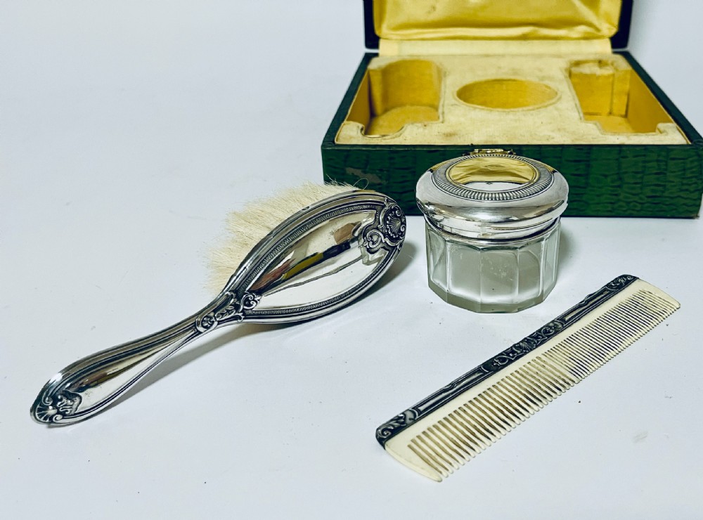 An open green textured box containing a French silver vanity set with a silver hairbrush, comb, and round lidded jar set into a yellow silk lining. View V