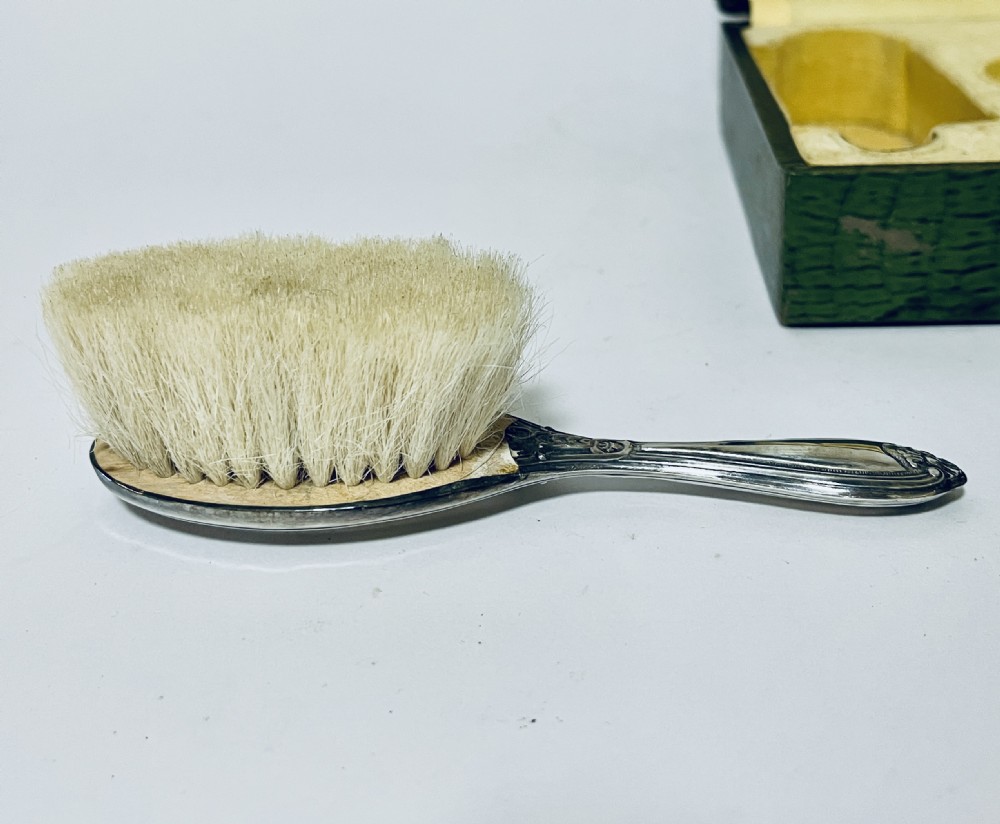 An open green textured box containing a French silver vanity set with a silver hairbrush, comb, and round lidded jar set into a yellow silk lining. View VII