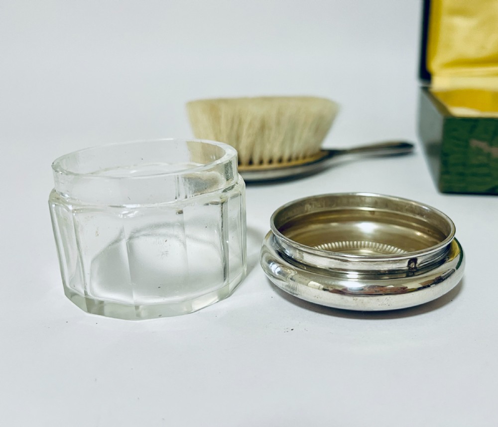 An open green textured box containing a French silver vanity set with a silver hairbrush, comb, and round lidded jar set into a yellow silk lining. View XV
