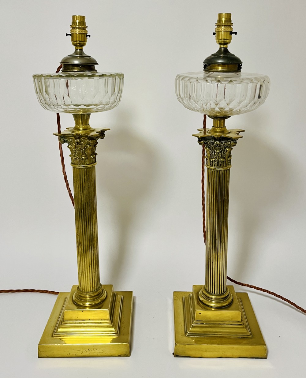 A pair of brass Corinthian column table lamps featuring stepped square plinths, fluted columns, acanthus capitals, and cut glass reservoirs.