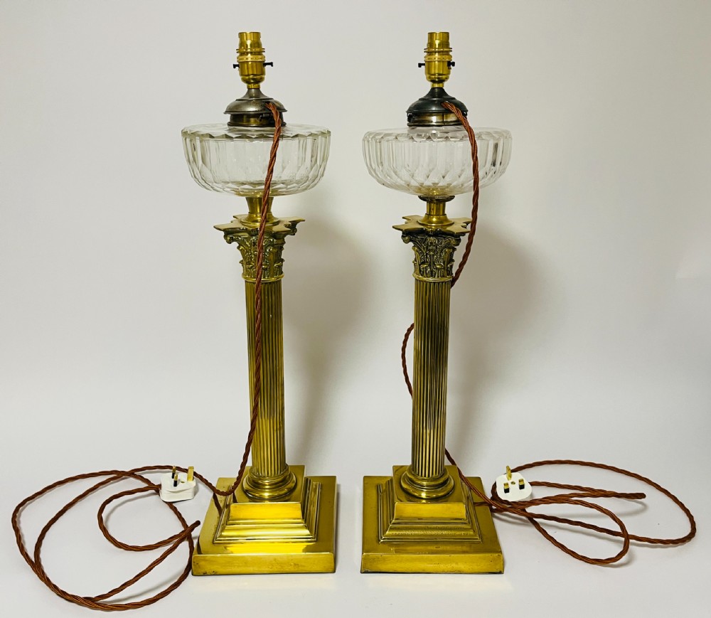 A pair of brass Corinthian column table lamps featuring stepped square plinths, fluted columns, acanthus capitals, and cut glass reservoirs. View VI