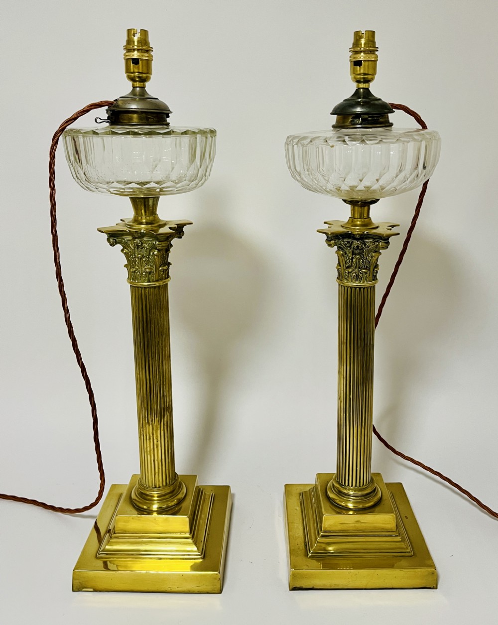 A pair of brass Corinthian column table lamps featuring stepped square plinths, fluted columns, acanthus capitals, and cut glass reservoirs. View IX