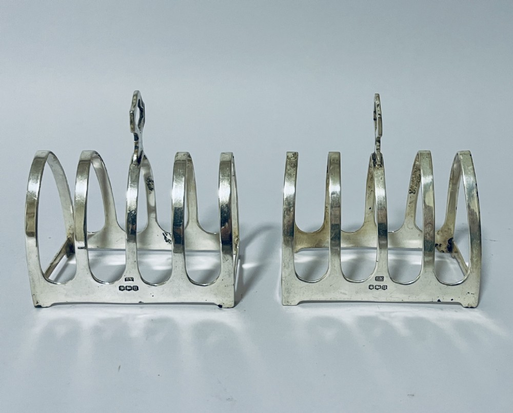 A pair of matching silver toast racks featuring five arched divisions, central geometric loop handles, and visible hallmarks along the front base rim.