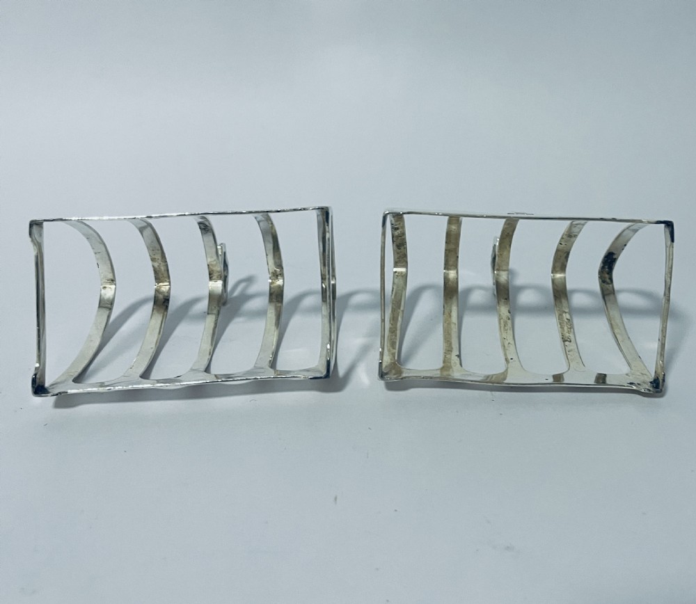 A pair of matching silver toast racks featuring five arched divisions, central geometric loop handles, and visible hallmarks along the front base rim. View VIII