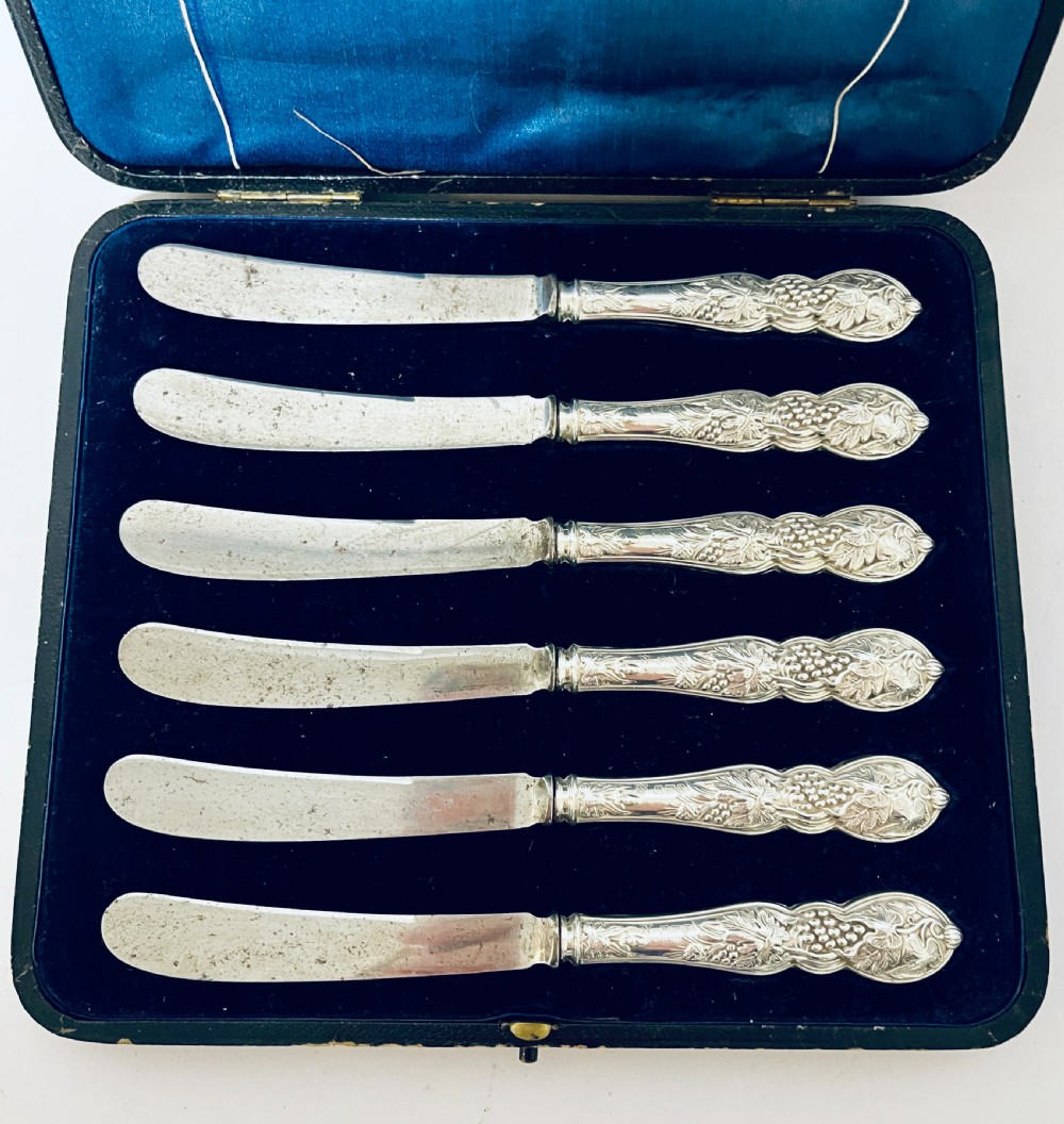 A set of six Victorian silver-handled butter knives with chased foliate handles, resting inside an open dark blue silk-lined presentation box. View II