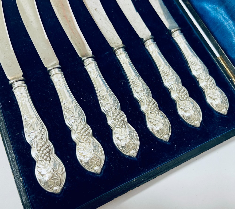 A set of six Victorian silver-handled butter knives with chased foliate handles, resting inside an open dark blue silk-lined presentation box. View III