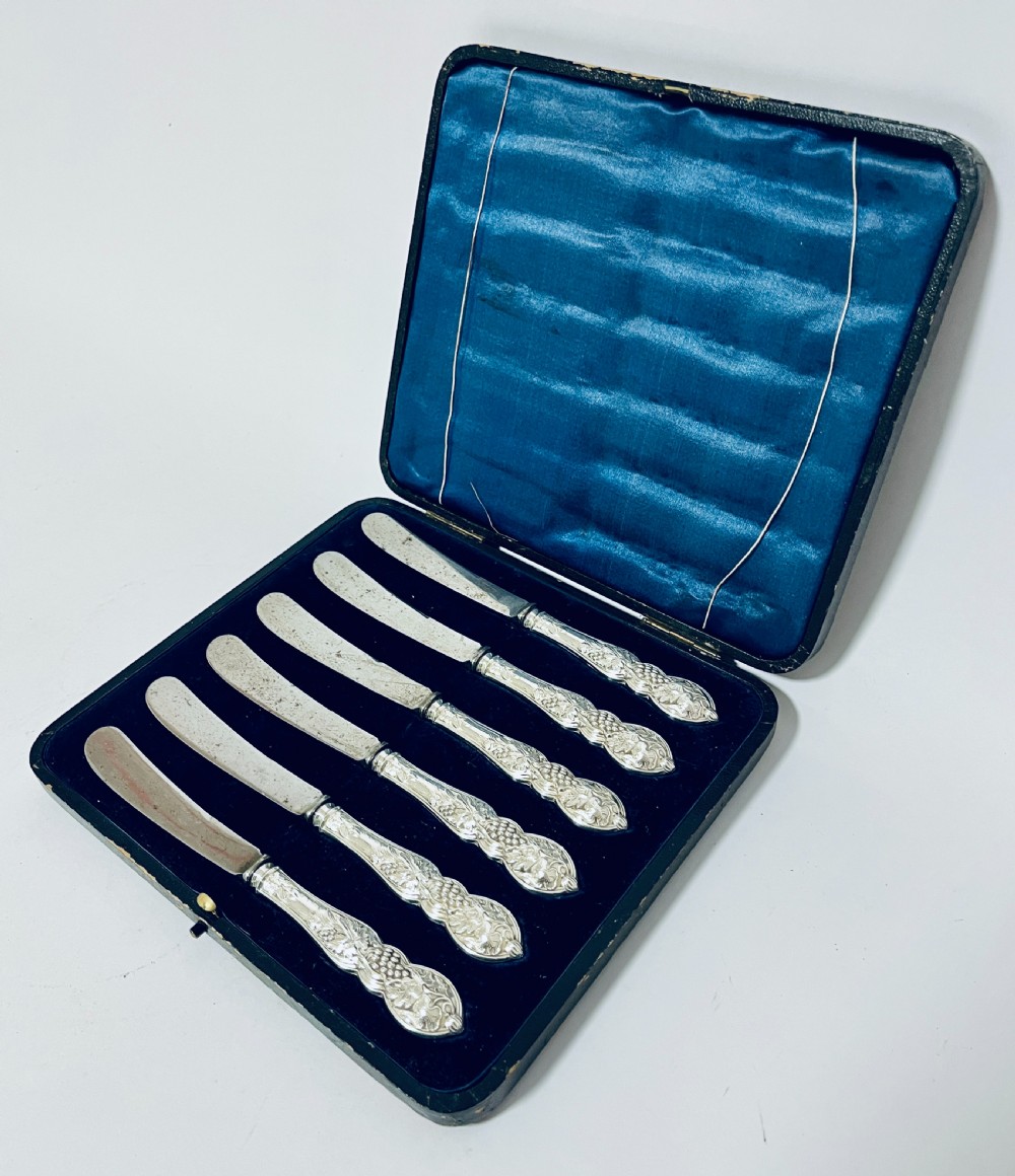 A set of six Victorian silver-handled butter knives with chased foliate handles, resting inside an open dark blue silk-lined presentation box. View IV