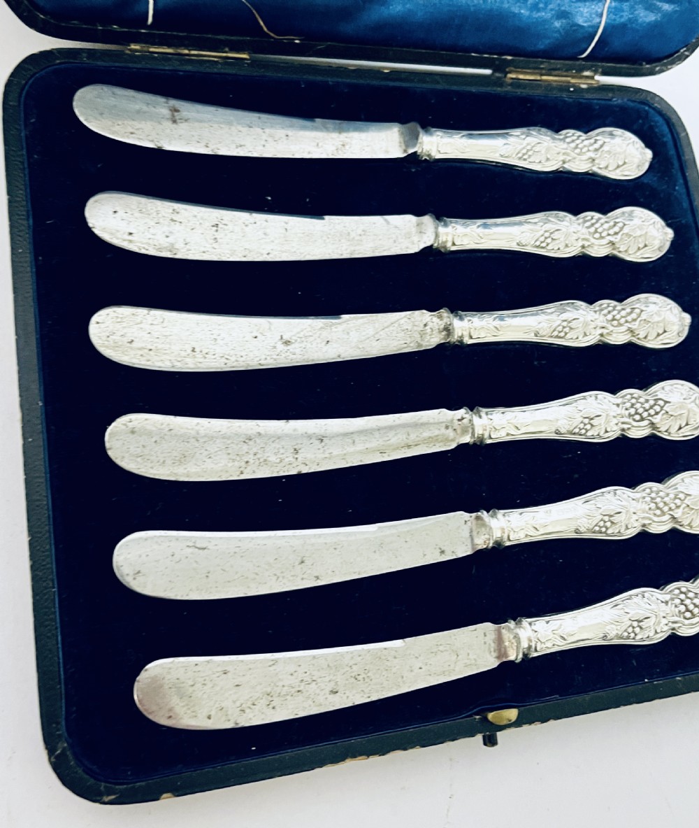 A set of six Victorian silver-handled butter knives with chased foliate handles, resting inside an open dark blue silk-lined presentation box. View XIII