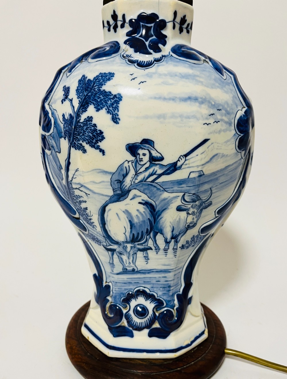 An 18th-century baluster-shaped Delftware ceramic vase decorated with blue and white pastoral scenes, mounted on a dark wooden plinth as a table lamp with brass fittings. View II