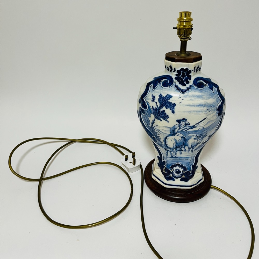 An 18th-century baluster-shaped Delftware ceramic vase decorated with blue and white pastoral scenes, mounted on a dark wooden plinth as a table lamp with brass fittings. View VI