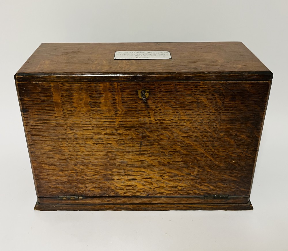 A rectangular Victorian dark oak stationery cabinet featuring a hinged drop-front, brass keyhole escutcheon, and an engraved metal plaque mounted on the top surface.