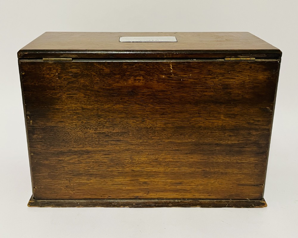 A rectangular Victorian dark oak stationery cabinet featuring a hinged drop-front, brass keyhole escutcheon, and an engraved metal plaque mounted on the top surface. View XI
