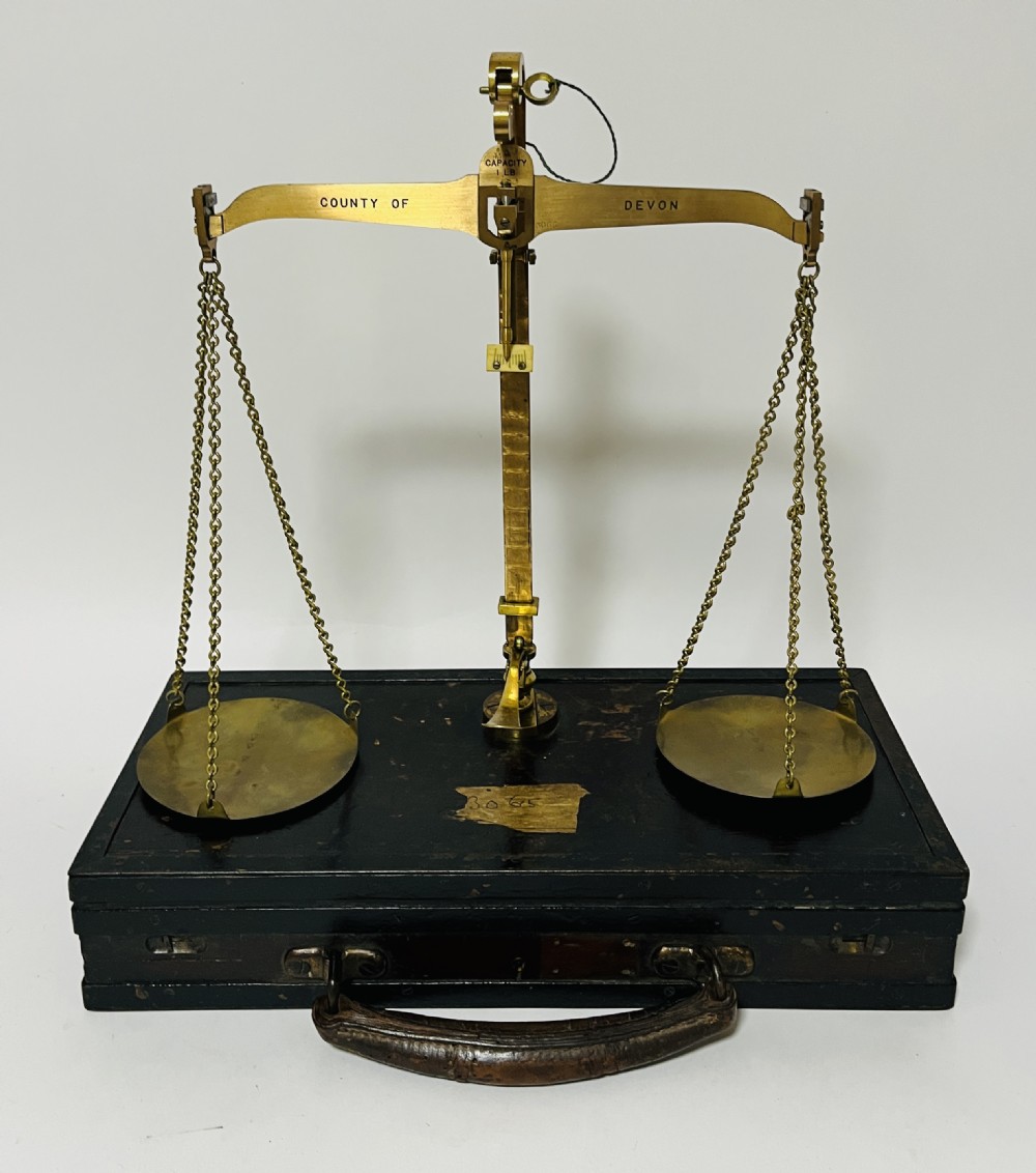 An antique brass balance scale stamped with County of Devon markings, resting on a black fitted transport case with two suspended brass pans.