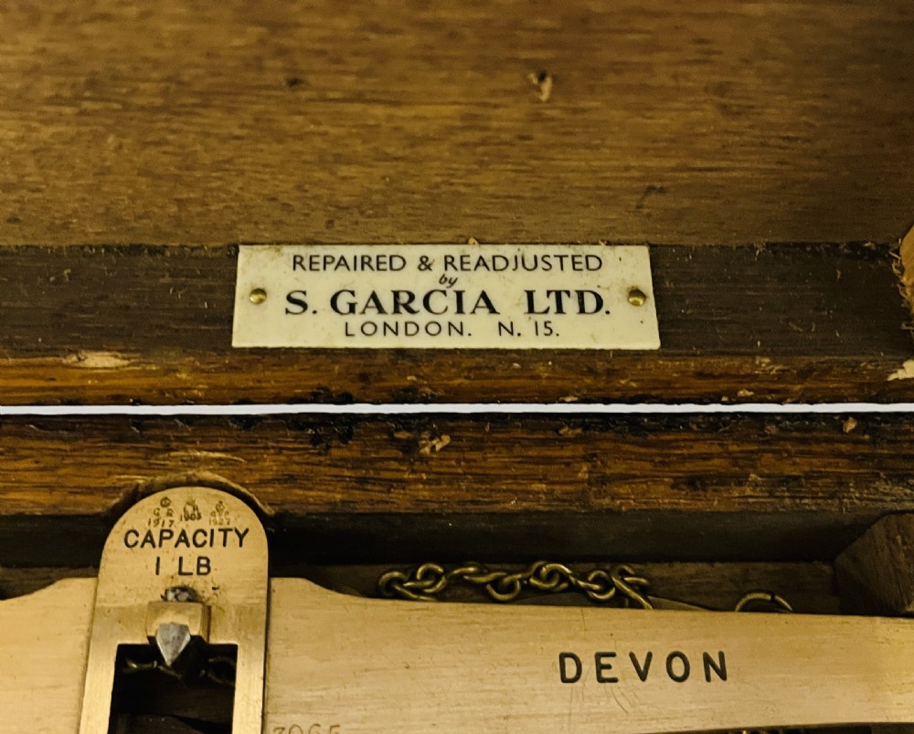 An antique brass balance scale stamped with County of Devon markings, resting on a black fitted transport case with two suspended brass pans. View IV