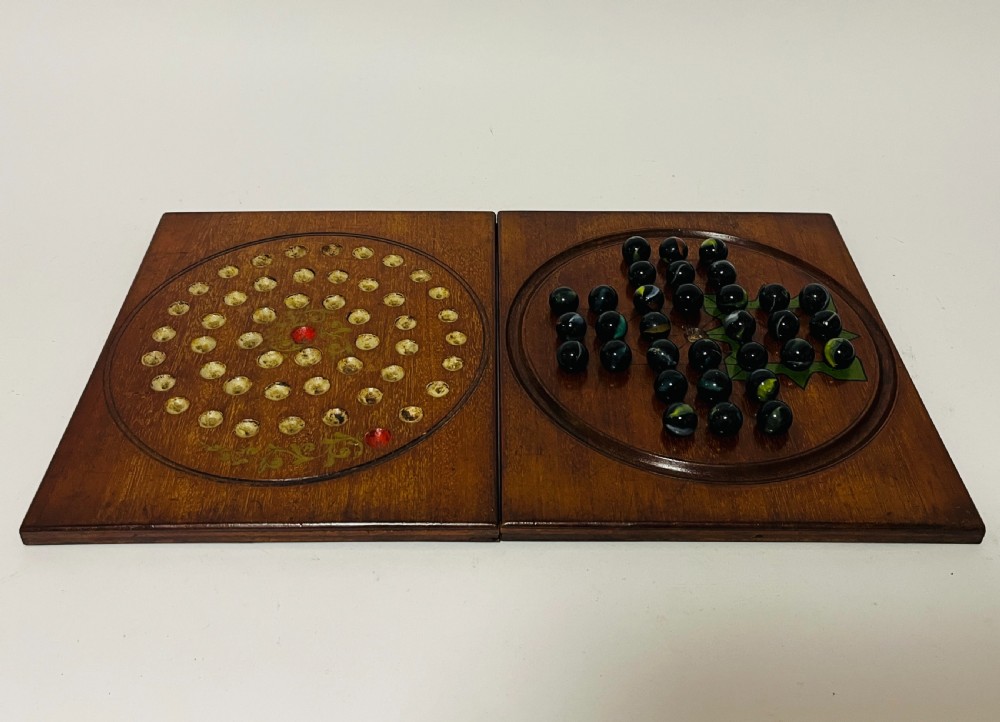 A two-panel antique mahogany solitaire games board featuring indented circular playing tracks alongside a collection of dark glass marbles.