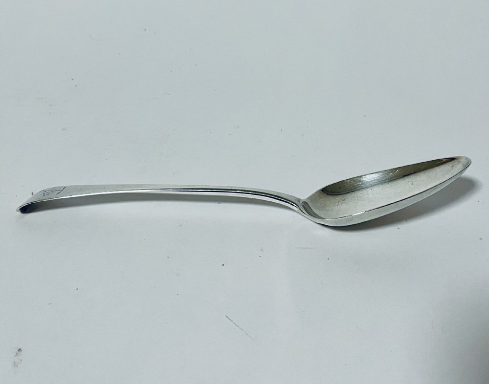 An 18th-century sterling silver serving spoon featuring a plain oval bowl and an engraved heraldic crest on the flat handle terminal. View IV