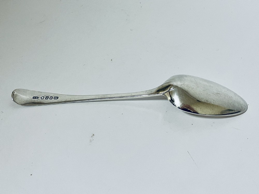 An 18th-century sterling silver serving spoon featuring a plain oval bowl and an engraved heraldic crest on the flat handle terminal. View VI