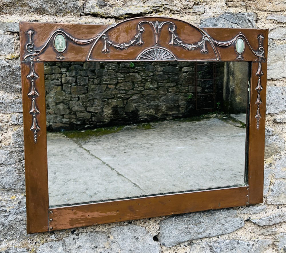 A rectangular Arts and Crafts wall mirror with an embossed copper frame featuring classical festoon motifs, an arched crest, and small green oval ceramic plaques.