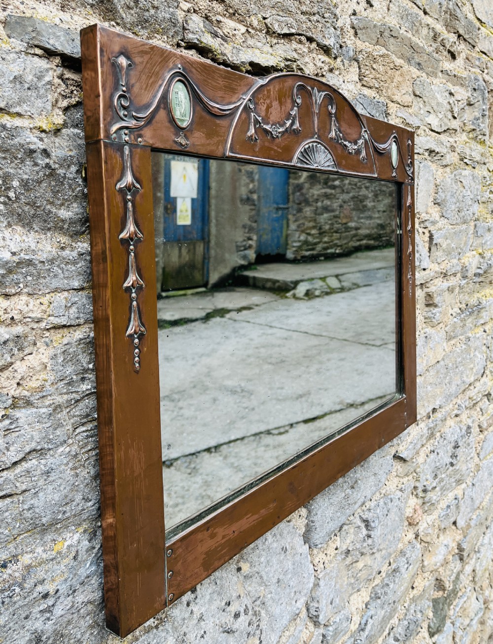 A rectangular Arts and Crafts wall mirror with an embossed copper frame featuring classical festoon motifs, an arched crest, and small green oval ceramic plaques. View VII