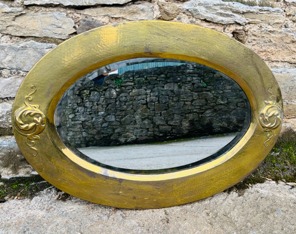 An oval wall mirror with a broad hammered brass frame featuring raised scroll motifs on the left and right sides.
