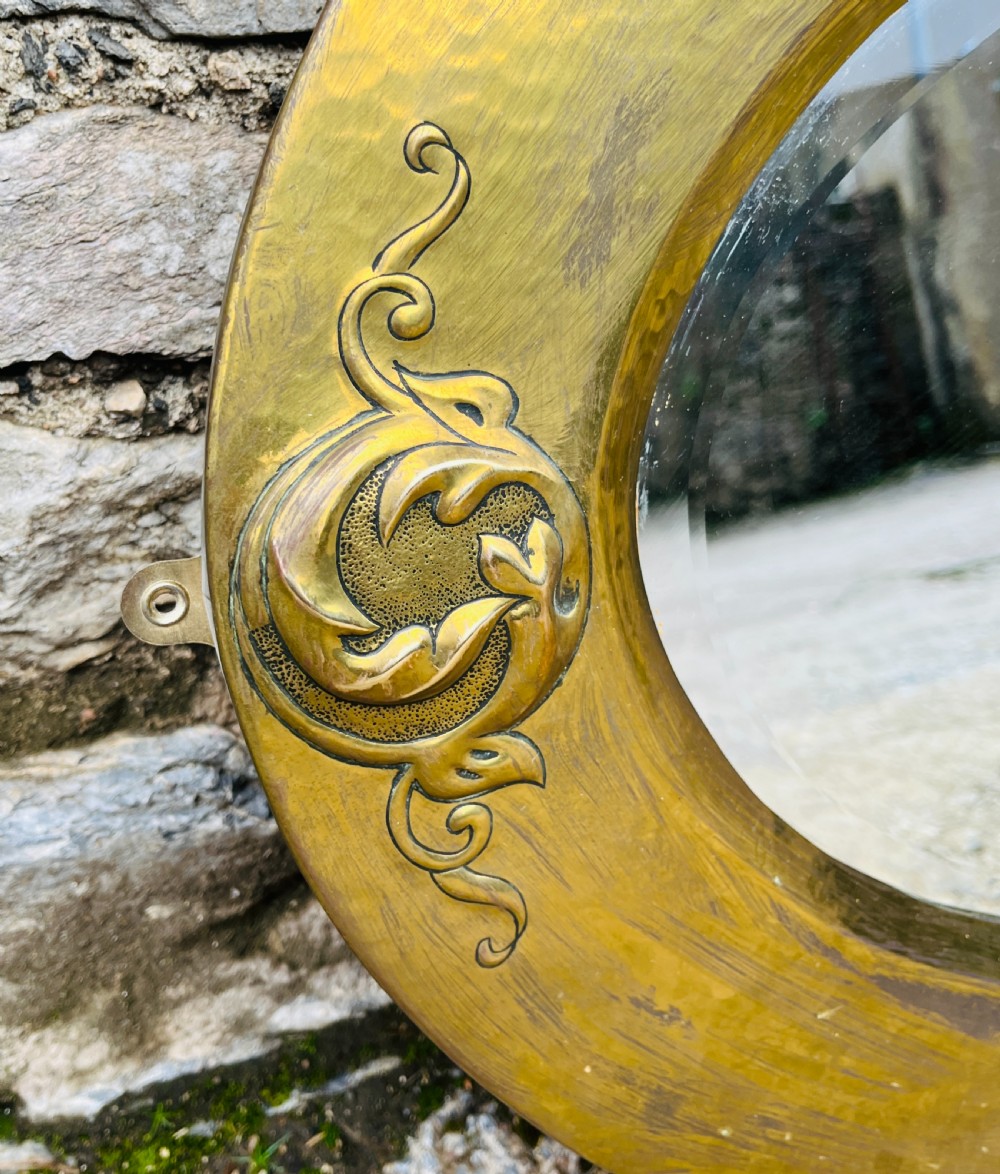 An oval wall mirror with a broad hammered brass frame featuring raised scroll motifs on the left and right sides. View II