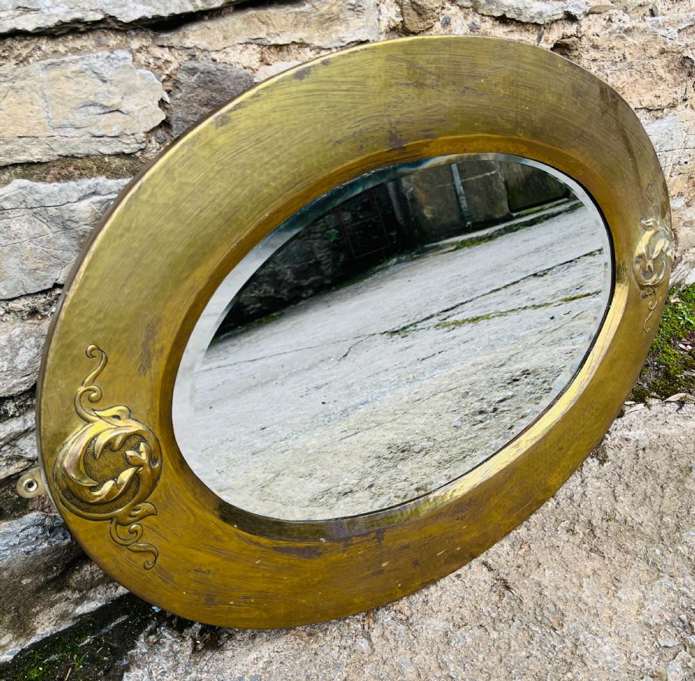 An oval wall mirror with a broad hammered brass frame featuring raised scroll motifs on the left and right sides. View III