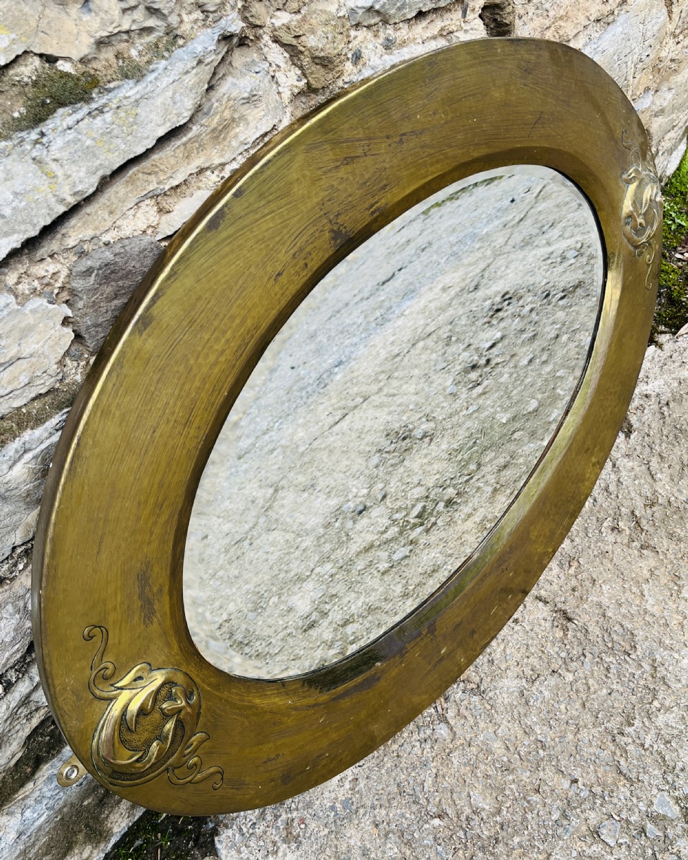 An oval wall mirror with a broad hammered brass frame featuring raised scroll motifs on the left and right sides. View IV