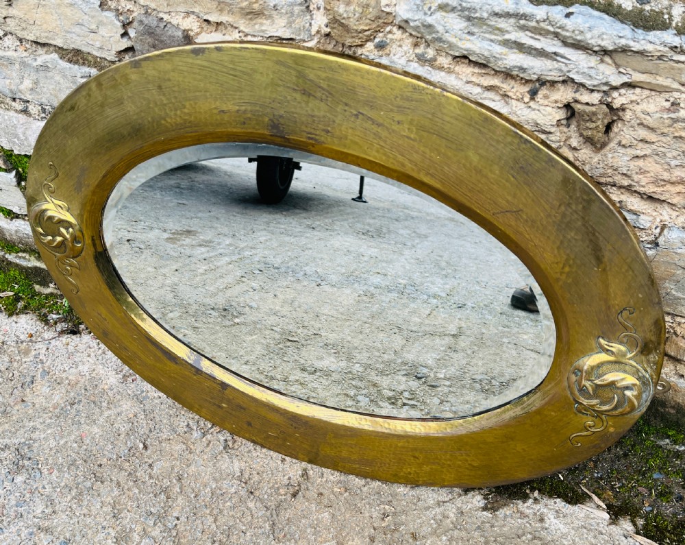 An oval wall mirror with a broad hammered brass frame featuring raised scroll motifs on the left and right sides. View V