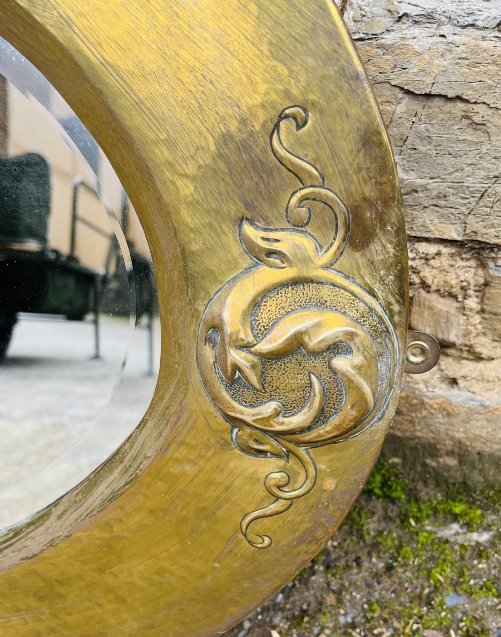 An oval wall mirror with a broad hammered brass frame featuring raised scroll motifs on the left and right sides. View VI
