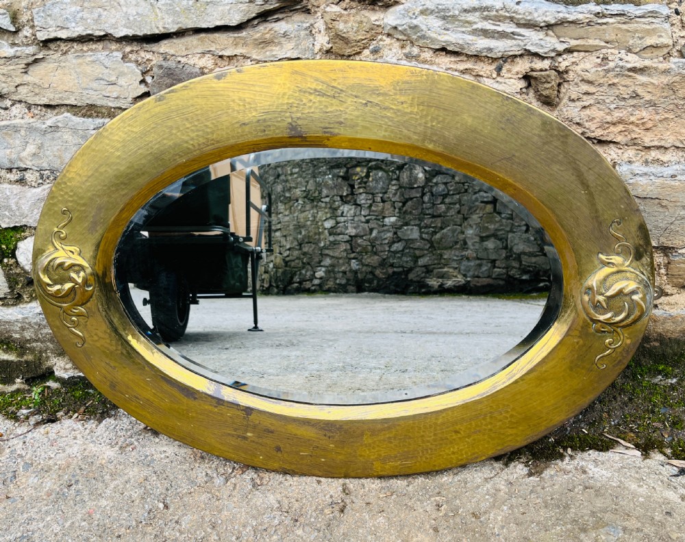 An oval wall mirror with a broad hammered brass frame featuring raised scroll motifs on the left and right sides. View VII