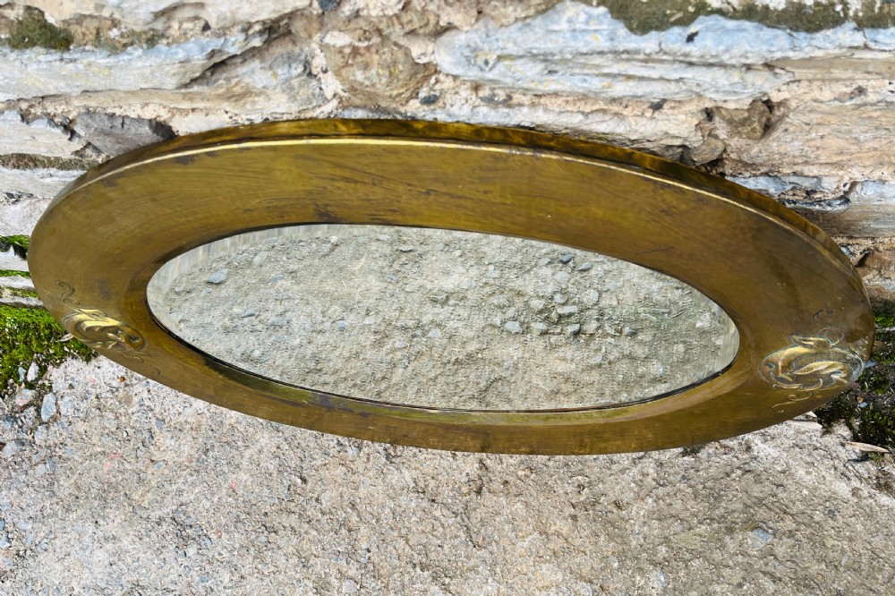 An oval wall mirror with a broad hammered brass frame featuring raised scroll motifs on the left and right sides. View VIII
