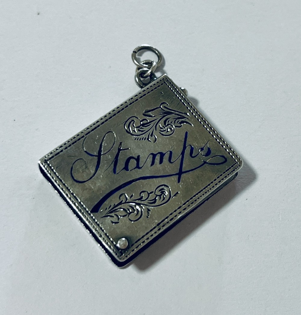 An antique silver stamp case featuring engraved foliate scrolls and script lettering with a suspension ring at the top.