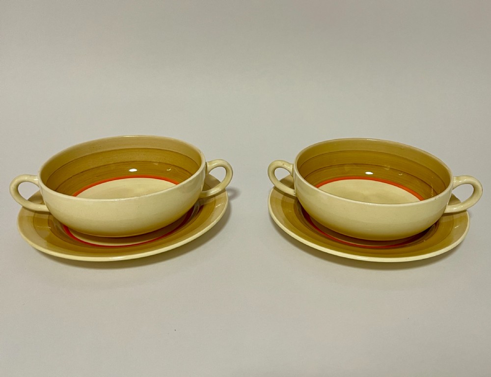A pair of cream glazed earthenware soup coupes with twin handles and matching saucers decorated with ochre yellow, brown, and orange horizontal bands.
