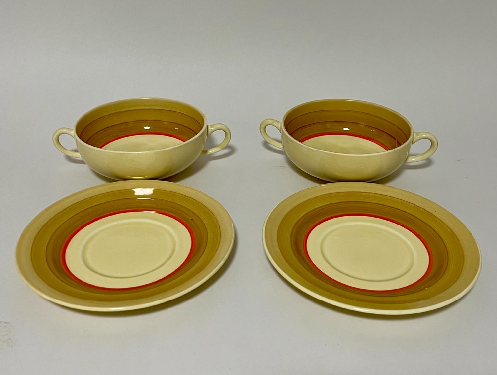 A pair of cream glazed earthenware soup coupes with twin handles and matching saucers decorated with ochre yellow, brown, and orange horizontal bands. View V