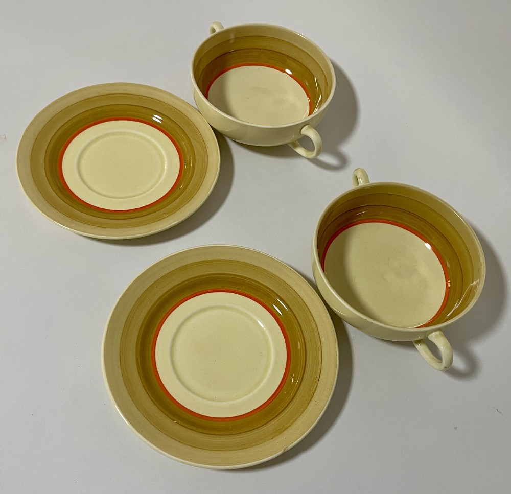 A pair of cream glazed earthenware soup coupes with twin handles and matching saucers decorated with ochre yellow, brown, and orange horizontal bands. View IX