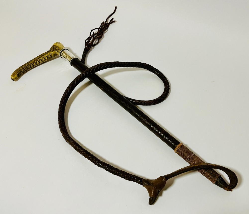 An antique hunting whip featuring a carved staghorn handle, a polished silver collar, a dark brown shaft, and a braided leather thong.