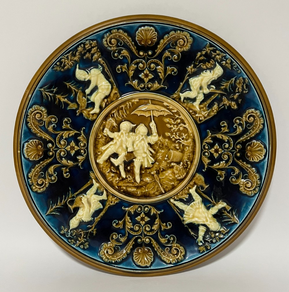 A circular Victorian majolica charger featuring relief figures of children and scrollwork in cobalt blue, ochre, and ivory glazes.