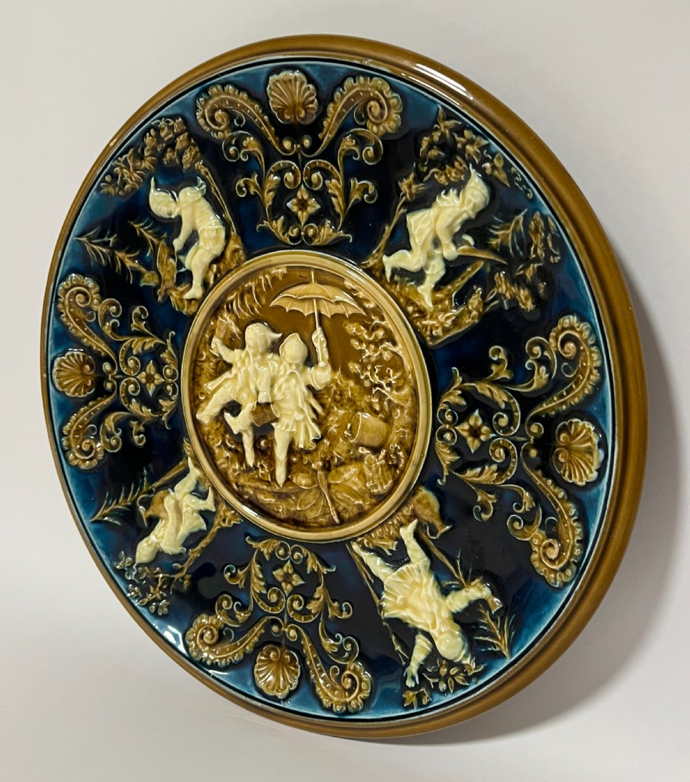 A circular Victorian majolica charger featuring relief figures of children and scrollwork in cobalt blue, ochre, and ivory glazes. View II