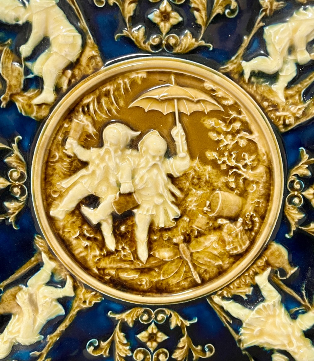 A circular Victorian majolica charger featuring relief figures of children and scrollwork in cobalt blue, ochre, and ivory glazes. View III