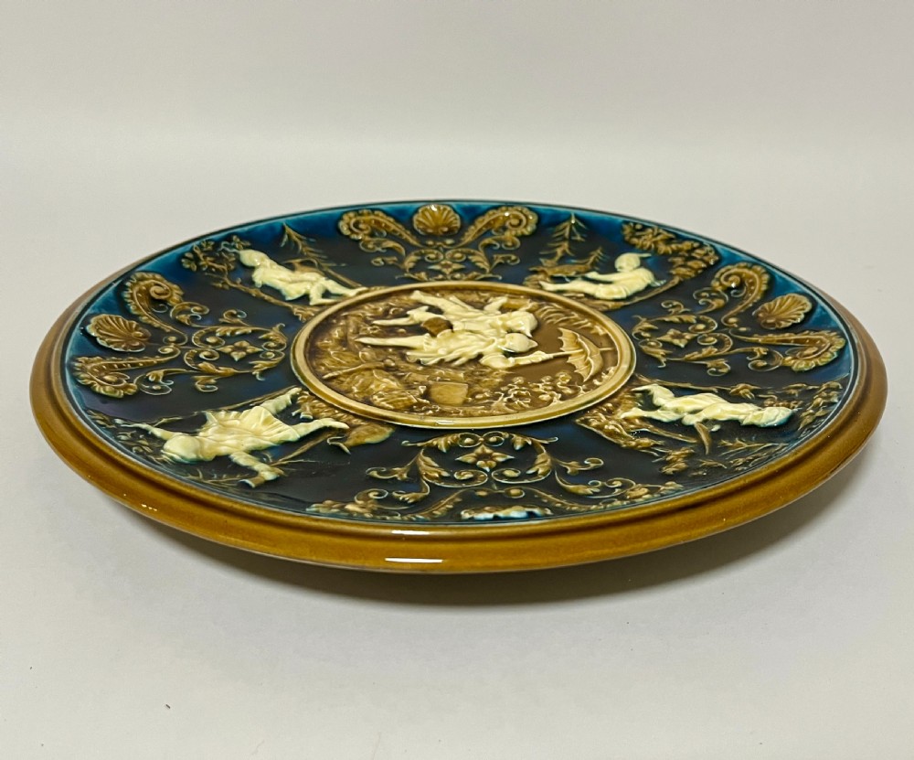 A circular Victorian majolica charger featuring relief figures of children and scrollwork in cobalt blue, ochre, and ivory glazes. View VIII